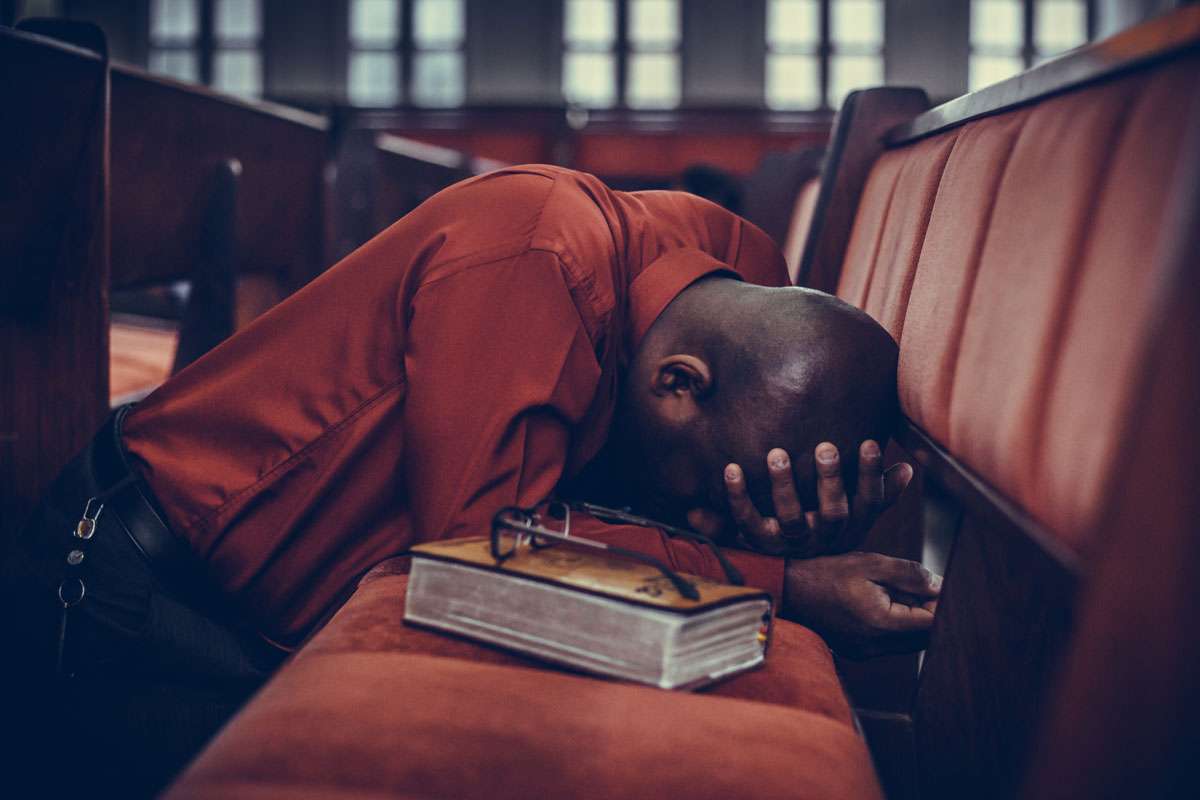 kneeling man forehead in hand head bent over resting on pew with bible and glasses in church